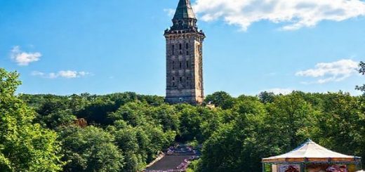 A majestic view of Hermannsdenkmal towering over the vibrant Teutoburg Forest, surrounded by lush summer greenery under a clear blue sky, with tourists enjoying outdoor activities and cultural events in the background, capturing the lively atmosphere of the season.