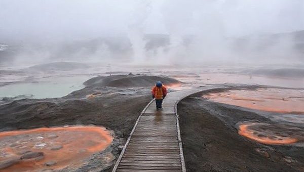 A surreal landscape at Craters of the Moon in winter, showcasing hissing steam vents and bubbling mud pools surrounded by a mystical mist. The scene contrasts the grey, chilly atmosphere with vibrant geothermal colors spiraling into the air, while a lone traveler in warm clothing walks along a misty boardwalk, evoking a sense of exploration and solitude in this otherworldly terrain.