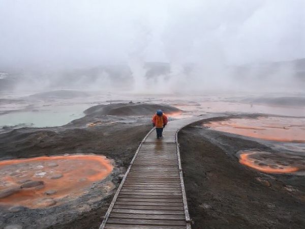A surreal landscape at Craters of the Moon in winter, showcasing hissing steam vents and bubbling mud pools surrounded by a mystical mist. The scene contrasts the grey, chilly atmosphere with vibrant geothermal colors spiraling into the air, while a lone traveler in warm clothing walks along a misty boardwalk, evoking a sense of exploration and solitude in this otherworldly terrain.