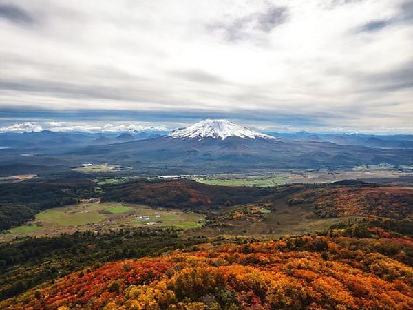 A breathtaking aerial view of the Ruapehu District, showcasing the majestic Mount Ruapehu surrounded by snow-capped peaks in winter, vibrant wildflowers in spring, golden autumn foliage, and lush greenery of summer, all under a dramatic sky that shifts from sunny to cloudy, capturing the essence of each season in this New Zealand paradise.