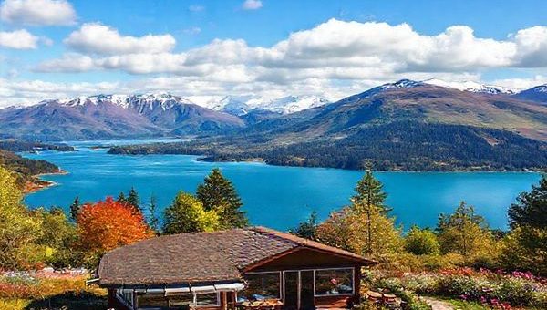 A stunning landscape of Taupō showcasing the vibrant blue of Lake Taupō surrounded by snow-capped mountains, lush greenery reflecting autumn colors, and wildflowers blooming in spring, with a cozy café in the foreground offering seasonal treats, under a bright blue sky dotted with fluffy white clouds.