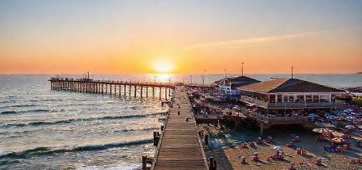 A majestic wooden pier stretching into the sparkling Baltic Sea, surrounded by vibrant summer crowds enjoying seafood at open-air restaurants, colorful beach umbrellas, and sunbathers along the sandy shore; in the distance, gentle waves lap against the pier as a warm sunset paints the sky in hues of orange and pink, creating a lively yet tranquil seaside atmosphere.