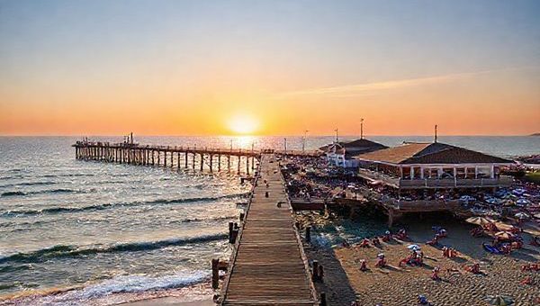 A majestic wooden pier stretching into the sparkling Baltic Sea, surrounded by vibrant summer crowds enjoying seafood at open-air restaurants, colorful beach umbrellas, and sunbathers along the sandy shore; in the distance, gentle waves lap against the pier as a warm sunset paints the sky in hues of orange and pink, creating a lively yet tranquil seaside atmosphere.