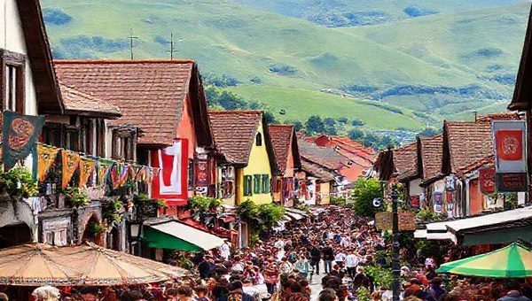 A vibrant Basque village bustling with life, showcasing local traditions: colorful festival banners, traditional dancers in folk costumes, a bustling market with artisanal food, and people gathered around enjoying Basque cuisine. In the background, rolling green hills and distinctive Basque architecture, with a subtle hint of the Euskara language visible on signposts and banners, creating an inviting atmosphere of cultural immersion.