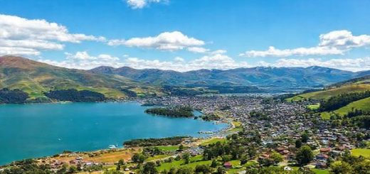 A breathtaking panoramic view of Ashburton District, New Zealand, capturing the vibrant green landscapes in summer with rolling hills and the Southern Alps in the background. Include serene Lake Hood bustling with water activities like boating and fishing, under a bright blue sky dotted with fluffy white clouds. Add charming small towns with colorful markets and lush parks filled with picnicking families. Render this scene in high definition, emphasizing the clarity and vividness of the summer colors.