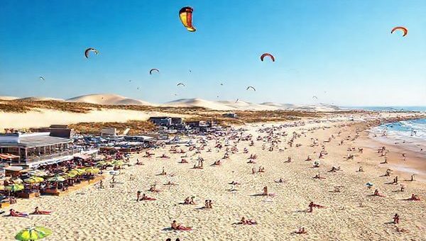 A breathtaking high-definition scene of Weststrand Sylt during a vibrant summer day, showcasing soft sandy beaches filled with sunbathers, colorful kites soaring overhead, and clear blue skies. In the background, capture the rolling dunes and stylish beachfront cafes bustling with visitors enjoying seafood and refreshing drinks. The atmosphere should exude warmth and joy, celebrating the lively summer spirit of this enchanting German island.