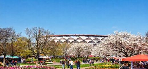 A picturesque scene of Gelsenkirchen in spring, showcasing vibrant flowers blooming in parks, bustling outdoor markets, and people enjoying leisurely strolls under clear blue skies. In the background, the iconic FC Schalke 04 stadium stands proudly, surrounded by blossoming trees. High definition.