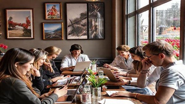 Create a high definition image of a cozy, inviting Dutch café scene where a diverse group of people, including travelers, culture enthusiasts, and business professionals, engage in learning Dutch. Show them using laptops, textbooks, and mobile apps, surrounded by Dutch artwork and traditional items. Incorporate elements like tulips and windmills visible through the café window, highlighting the rich cultural heritage of the Netherlands.