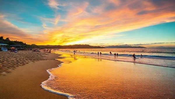 **High definition image prompt:** A serene view of Raumati Beach on New Zealand's Kapiti Coast, showcasing golden sands and lush greenery under a vibrant sunset sky. In the foreground, gentle waves lap at the shore, while in the background, families enjoy beach sports and paddleboarding. Include colorful beach umbrellas and a bustling outdoor cafe scene, capturing the lively yet laid-back summer atmosphere. Add hints of local flora and the silhouette of a distant mountain range.