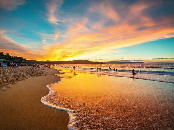 **High definition image prompt:** A serene view of Raumati Beach on New Zealand's Kapiti Coast, showcasing golden sands and lush greenery under a vibrant sunset sky. In the foreground, gentle waves lap at the shore, while in the background, families enjoy beach sports and paddleboarding. Include colorful beach umbrellas and a bustling outdoor cafe scene, capturing the lively yet laid-back summer atmosphere. Add hints of local flora and the silhouette of a distant mountain range.