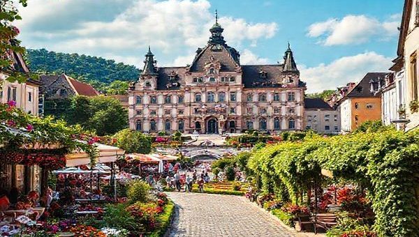 A high-definition image of Karlsruhe's picturesque cityscape, showcasing the Karlsruhe Palace surrounded by vibrant summer gardens filled with blooming flowers and lively people enjoying outdoor cafes. The scene captures the essence of a sunny day, with festival decorations and warm colors reflecting the vibrant atmosphere of the city. Include elements of the Black Forest in the background, with vines and greenery along the cobblestone streets, to highlight the blend of history and nature.