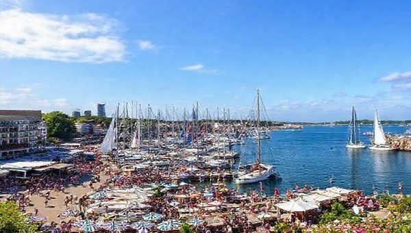 A high-definition image of Kiel, Germany, captured in summer during the vibrant Kiel Week festival, showcasing bustling harbor activity with colorful sailboats racing, lively crowds enjoying music and food, and the picturesque skyline adorned with lush greenery and blooming flowers. Include glimpses of the beautiful coastline, such as Schönberger Strand, and the festive atmosphere of outdoor events under a blue sky.