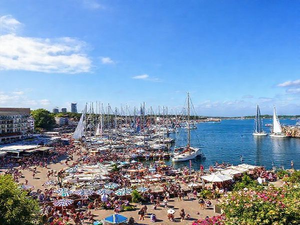 A high-definition image of Kiel, Germany, captured in summer during the vibrant Kiel Week festival, showcasing bustling harbor activity with colorful sailboats racing, lively crowds enjoying music and food, and the picturesque skyline adorned with lush greenery and blooming flowers. Include glimpses of the beautiful coastline, such as Schönberger Strand, and the festive atmosphere of outdoor events under a blue sky.