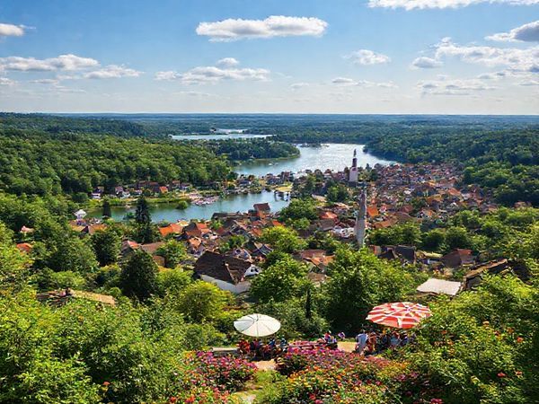 A stunning view of Landkreis Potsdam-Mittelmark in summer, showcasing vibrant greenery and a serene lake glittering under the sun. Add charming small towns in the foreground with locals enjoying outdoor festivities, surrounded by lush forests and colorful flowers. The scene is filled with warmth and the essence of German culture, highlighting outdoor activities like kayaking and cycling. Please create this image in high definition.