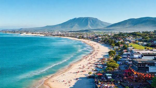 A stunning aerial view of Tauranga, New Zealand, showcasing its vibrant coastline with white sandy beaches, the imposing Mount Maunganui in the background, and lush green vineyards, under a clear blue sky. The scene captures the lively atmosphere of summer with sunbathers on the beach, vibrant cafes spilling onto the streets, and colorful lanterns illuminating a summer festival. Render in high definition for rich detail and clarity.