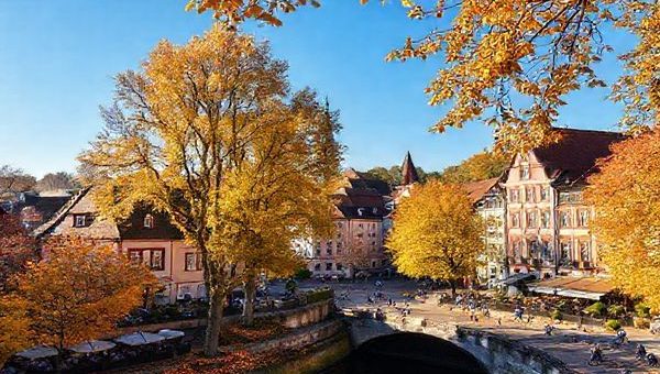 A picturesque view of Gießen in autumn, showcasing vibrant trees in hues of gold and amber surrounding charming historic buildings. Include a lively university atmosphere with students enjoying outdoor cafes, and a clear blue sky. Capture the essence of fall with scattered leaves on the ground and people cycling along the Lahn river. Make this image in high definition.