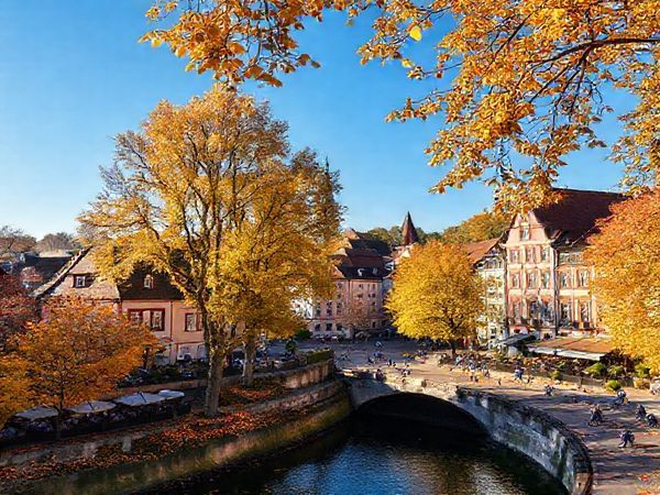 A picturesque view of Gießen in autumn, showcasing vibrant trees in hues of gold and amber surrounding charming historic buildings. Include a lively university atmosphere with students enjoying outdoor cafes, and a clear blue sky. Capture the essence of fall with scattered leaves on the ground and people cycling along the Lahn river. Make this image in high definition.