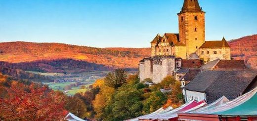 A high-definition image capturing the enchanting landscapes of Niederbayern in autumn, showcasing vibrant golds, reds, and browns among rolling hills, historic landmarks like Trausnitz Castle, and bustling harvest festivals, with locals joyfully participating in outdoor markets under a clear blue sky.