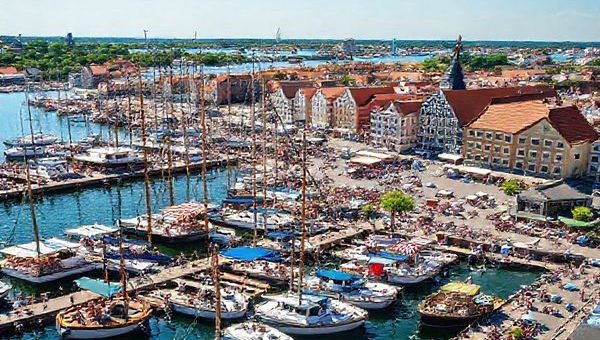 Create a high definition image depicting the vibrant port city of Bremerhaven in summer. Show bustling docks filled with tourists enjoying seafood festivals, yachts sailing through the lively harbor, and the German Maritime Museum under the warm sun. Highlight the bright colors of the blue sea, lush greenery of parks, and the festive atmosphere created by locals and visitors. Capture the maritime charm and innovation that define this charming North Sea coastal city.