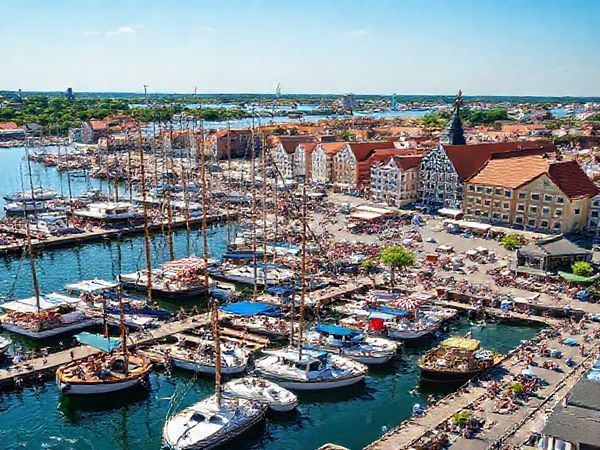 Create a high definition image depicting the vibrant port city of Bremerhaven in summer. Show bustling docks filled with tourists enjoying seafood festivals, yachts sailing through the lively harbor, and the German Maritime Museum under the warm sun. Highlight the bright colors of the blue sea, lush greenery of parks, and the festive atmosphere created by locals and visitors. Capture the maritime charm and innovation that define this charming North Sea coastal city.