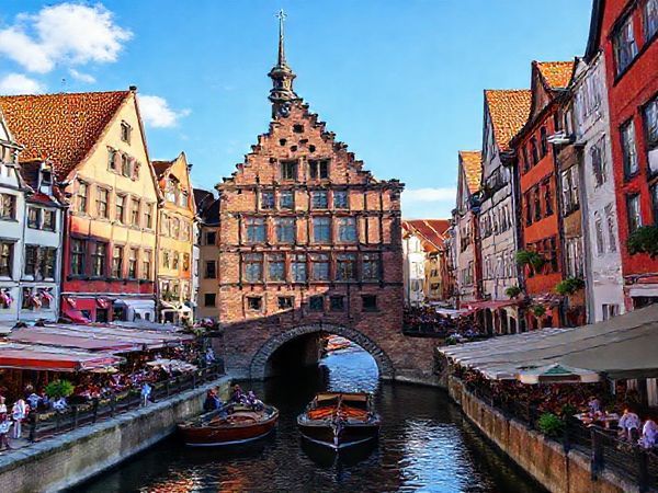 Create a high definition image of Lübeck Old Town in summer, showcasing its medieval architecture with vibrant colors and bustling market squares. Include the iconic Holsten Gate with sunlit canals, lively outdoor cafes, and people enjoying a boat tour along the Trave River. Capture the joyous atmosphere of the Lübeck Cultural Festival, with music and art blending into the charming cobbled streets lined with gabled houses under a clear blue sky.