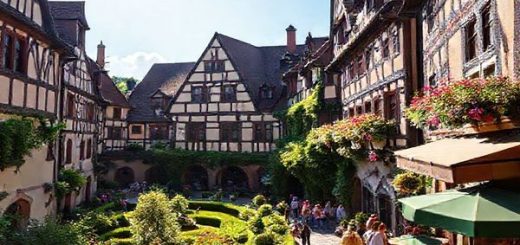 "Create a high-definition image of the Fuggerei, showcasing its charming Gothic and Renaissance architecture during a vibrant summer day. Capture lush gardens in full bloom, quaint pathways filled with visitors admiring the historic buildings, and the warm sun illuminating the scene. Include the inviting ambiance of the Michlstube beer garden nearby, filled with people enjoying Bavarian brews amidst the beauty of this serene enclave in Augsburg, Germany."