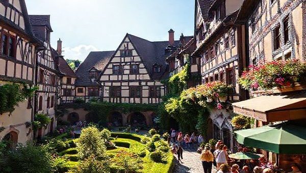 "Create a high-definition image of the Fuggerei, showcasing its charming Gothic and Renaissance architecture during a vibrant summer day. Capture lush gardens in full bloom, quaint pathways filled with visitors admiring the historic buildings, and the warm sun illuminating the scene. Include the inviting ambiance of the Michlstube beer garden nearby, filled with people enjoying Bavarian brews amidst the beauty of this serene enclave in Augsburg, Germany."