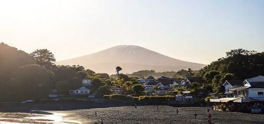 A serene coastal village scene of Ōakura, New Zealand, showcasing its stunning black sand beaches and lush greenery. Include the majestic Mount Taranaki in the background under a bright, sunlit sky, with a few surfers riding gentle waves. Add a picturesque sunset in the distance, casting vibrant hues of orange and pink. The foreground features locals enjoying a leisurely beach day, along with hints of the village’s charming cafes and cultural events. Create this image in high definition.