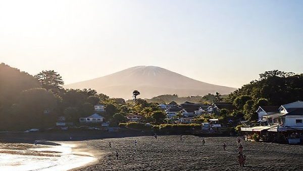 A serene coastal village scene of Ōakura, New Zealand, showcasing its stunning black sand beaches and lush greenery. Include the majestic Mount Taranaki in the background under a bright, sunlit sky, with a few surfers riding gentle waves. Add a picturesque sunset in the distance, casting vibrant hues of orange and pink. The foreground features locals enjoying a leisurely beach day, along with hints of the village’s charming cafes and cultural events. Create this image in high definition.
