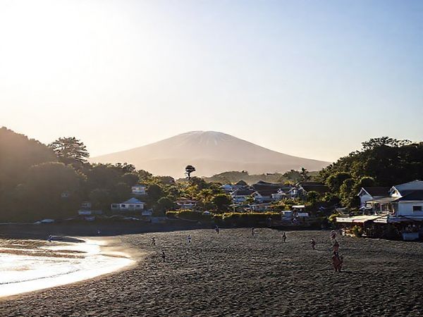 A serene coastal village scene of Ōakura, New Zealand, showcasing its stunning black sand beaches and lush greenery. Include the majestic Mount Taranaki in the background under a bright, sunlit sky, with a few surfers riding gentle waves. Add a picturesque sunset in the distance, casting vibrant hues of orange and pink. The foreground features locals enjoying a leisurely beach day, along with hints of the village’s charming cafes and cultural events. Create this image in high definition.