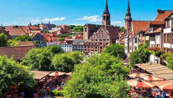Create a high-definition image of Nürnberg in summer, showcasing vibrant outdoor scenes with lush green parks, lively beer gardens filled with people clinking mugs, and a backdrop of medieval architecture under a clear blue sky. Include colorful street festivals with artists and musicians, capturing the city's energetic atmosphere and the essence of Bavarian culture in full bloom.