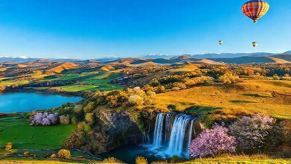 A vibrant scene showcasing the breathtaking landscapes of Waikato District in New Zealand, highlighting the rolling hills, lush pastures, and a majestic waterfall under a deep blue sky. Incorporate cultural elements such as a Maori cultural performance in the foreground and the Waikato Cherry Blossom Festival in full bloom nearby. Include various outdoor activities like kayaking on a serene lake and hot air balloons drifting in the sky during fall. Capture the essence of all four seasons, emphasizing the contrast of summer's warmth, winter's tranquility, autumn's vibrant colors, and spring's lively blossoms, all rendered in high definition.