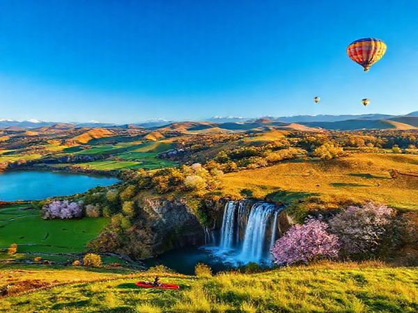 A vibrant scene showcasing the breathtaking landscapes of Waikato District in New Zealand, highlighting the rolling hills, lush pastures, and a majestic waterfall under a deep blue sky. Incorporate cultural elements such as a Maori cultural performance in the foreground and the Waikato Cherry Blossom Festival in full bloom nearby. Include various outdoor activities like kayaking on a serene lake and hot air balloons drifting in the sky during fall. Capture the essence of all four seasons, emphasizing the contrast of summer's warmth, winter's tranquility, autumn's vibrant colors, and spring's lively blossoms, all rendered in high definition.