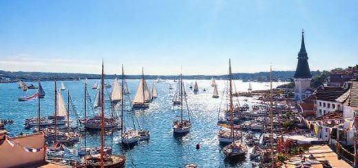 A picturesque view of Flensburg Harbour during summer, filled with vibrant sailing boats gliding across the sparkling fjord under a bright blue sky. The charming waterfront is alive with bustling cafes and people dining alfresco, enjoying fresh seafood while basking in the golden sunlight. The scene captures the lively atmosphere of the Flensburg Rum Regatta, with colorful flags fluttering and festive decorations adorning the waterfront. Render this in high definition, emphasizing the lively colors and intricate details of the boats and the picturesque harbor.