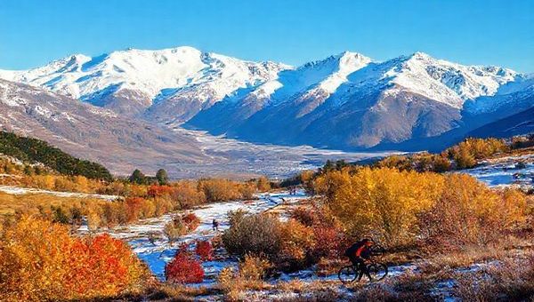 A breathtaking high-definition landscape of New Zealand's Canterbury Region, showcasing the majestic Southern Alps covered in snow during winter, contrasting with vibrant autumn colors in the foreground. Include elements of outdoor adventure like skiers on the slopes and cyclists exploring serene trails, all under a clear blue sky. The image should encapsulate the diverse beauty of Canterbury across seasons, highlighting its rich natural scenery and inviting atmosphere.