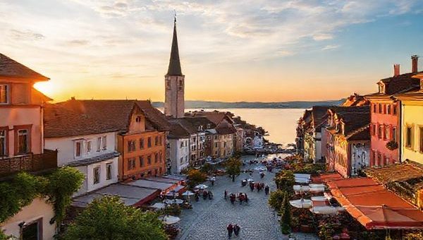 A serene scene of Konstanz by Lake Constance, depicting the charming Old Town with medieval architecture, vibrant cafés spilling onto cobbled streets, and lively festival celebrations in the summer. Include the lake reflecting the warm sunset, locals enjoying sailing and windsurfing, surrounded by lush greenery and blooming flowers in spring, contrasted with autumn foliage in warm hues. Capture the essence of a cultural hub, with hints of winter's Christmas market ambience. Create this image in high definition.