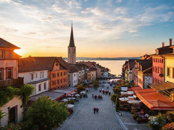 A serene scene of Konstanz by Lake Constance, depicting the charming Old Town with medieval architecture, vibrant cafés spilling onto cobbled streets, and lively festival celebrations in the summer. Include the lake reflecting the warm sunset, locals enjoying sailing and windsurfing, surrounded by lush greenery and blooming flowers in spring, contrasted with autumn foliage in warm hues. Capture the essence of a cultural hub, with hints of winter's Christmas market ambience. Create this image in high definition.