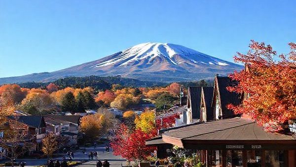 A stunning landscape of Stratford, New Zealand, showcasing Mount Taranaki against a vibrant autumn backdrop of golden and crimson leaves. Capture the serene ambiance of the town with quaint local shops and blooming gardens, while people leisurely stroll along scenic paths. The sky is a clear blue, hinting at mild temperatures, and a cozy café sits invitingly in the foreground. High definition.