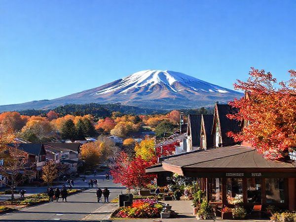 A stunning landscape of Stratford, New Zealand, showcasing Mount Taranaki against a vibrant autumn backdrop of golden and crimson leaves. Capture the serene ambiance of the town with quaint local shops and blooming gardens, while people leisurely stroll along scenic paths. The sky is a clear blue, hinting at mild temperatures, and a cozy café sits invitingly in the foreground. High definition.