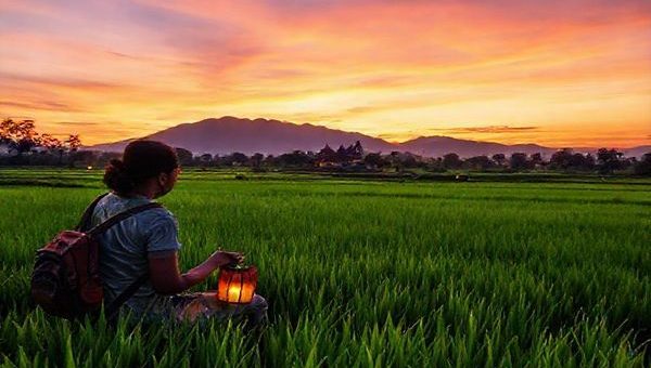 A serene Lao landscape at dusk, featuring lush green rice paddies and a distant temple, where a traveler practices Lao with a friendly local. The sky is painted in hues of orange and purple, reflecting the sunset, while soft lantern lights illuminate the scene. Captured in high definition, emphasizing the beauty of cultural exchange and the joy of learning a new language amidst nature.