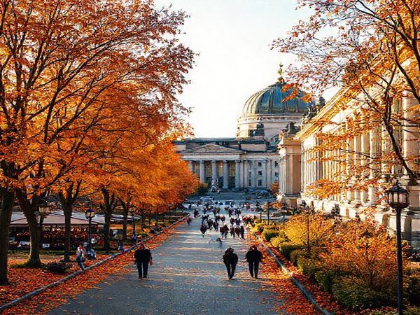 Create a high definition image of Museum Island in Berlin during the vibrant autumn season, showcasing the iconic museums surrounded by trees adorned in brilliant shades of gold, orange, and red. Capture a gentle path lined with fallen leaves, with visitors leisurely strolling, mingling with lively street performers and enjoying outdoor cafes. The warm, soft light of the fall sun should illuminate the grand architecture, creating a picturesque, inviting atmosphere that reflects the rich cultural essence of this UNESCO World Heritage Site.