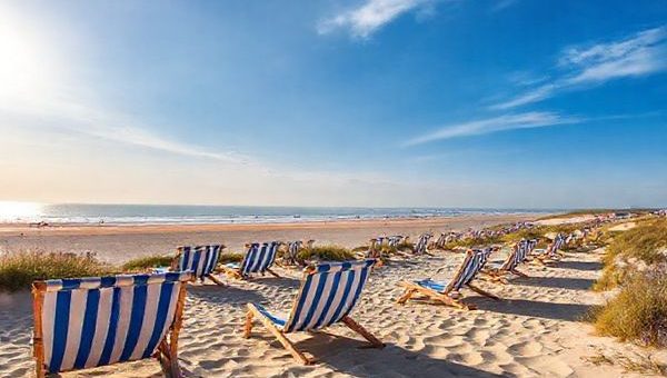 A breathtaking high-definition view of Sylt, Germany, capturing the essence of all four seasons. The image should feature the stunning sandy beaches with iconic striped beach chairs under a bright summer sun, transitioning to serene winter landscapes with frosty dunes and dramatic skies. Add vibrant autumn heather in hues of purple and charming cafes bustling with activity, alongside blooming spring flowers and cyclists in the fresh sea breeze. The scene should seamlessly blend these seasonal elements, radiating the relaxed luxury of this picturesque North Sea island.