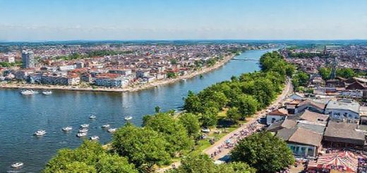 A panoramic high-definition view of Ludwigshafen am Rhein during a vibrant summer day, showcasing the bustling Rhine waterfront filled with families enjoying boat rides, parks in full bloom, and the festive atmosphere of the Rheinschanzenfest with colorful stalls and joyful crowds. The cityscape blends modern architecture with historical buildings against a backdrop of clear blue skies and lush green landscapes.