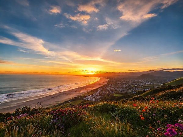 A breathtaking sunset over the Kāpiti Coast District, showcasing golden beaches and lush green hills. Include vibrant summer activity scenes with surfers riding the waves, families picnicking on the shore, and colorful flowers blooming in the nearby gardens. Capture charming towns in the background and a serene, inviting sky filled with warm hues. Please make the image in high definition.