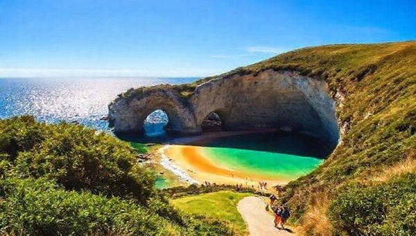 A breathtaking view of Cathedral Cove in New Zealand, showcasing the stunning limestone archways framed by azure waters and golden sands under a vibrant blue sky. Lush greenery lines the winding path leading to the cove, with a few tourists enjoying the serene atmosphere. The scene captures the essence of summer, with sunshine glistening on the ocean surface, inviting the viewer to bask in this coastal paradise. Please create this image in high definition.