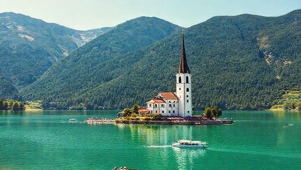 A picturesque scene of Königssee in summer, showcasing its serene emerald waters surrounded by vibrant green mountains under a bright blue sky. Capture the iconic St. Bartholomä pilgrimage church on the lake's shores, with visitors enjoying boat rides and hiking trails. The landscape should be alive with summer activity and vivid colors, emphasizing tranquility alongside lively outdoor pursuits. Ensure the image is in high definition.