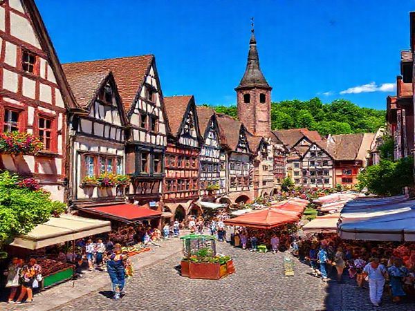 "Create a high definition image of Freiburg, Germany in the heart of summer, showcasing cobbled streets bustling with locals and tourists, vibrant markets filled with colorful stalls, and lush greenery surrounding the city. The scene should capture the warmth of the sun, the atmosphere of lively festivals, and the picturesque architecture of the medieval old town, all under a clear blue sky."