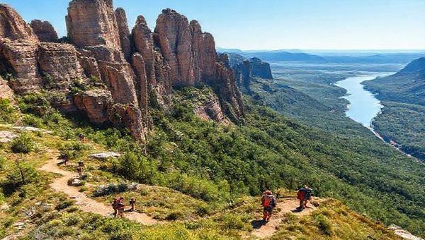 A breathtaking high-definition landscape of the Elbe Sandstone Mountains, showcasing the dramatic rock formations adorned with vibrant summer greenery and the glistening Elbe River in the background. Include hikers on the trails enjoying the outdoor adventure, with a clear blue sky overhead.