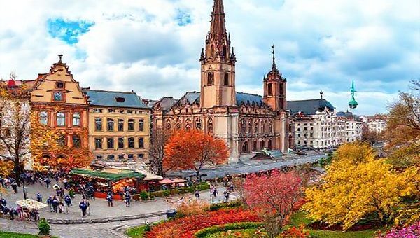 "Create a high-definition image of Leipzig during each season: a vibrant summer scene with bustling outdoor festivals and musicians, a cozy winter wonderland adorned with twinkling lights and a Christmas market, a picturesque autumn landscape with amber and golden leaves, and a blooming spring garden filled with colorful blossoms. Blend these four seasonal elements into one panoramic view of the enchanting city of Leipzig."