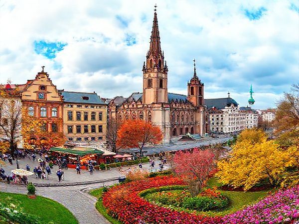 "Create a high-definition image of Leipzig during each season: a vibrant summer scene with bustling outdoor festivals and musicians, a cozy winter wonderland adorned with twinkling lights and a Christmas market, a picturesque autumn landscape with amber and golden leaves, and a blooming spring garden filled with colorful blossoms. Blend these four seasonal elements into one panoramic view of the enchanting city of Leipzig."